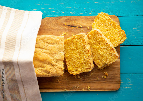 Cornbread is bread containing cornmeal, corn flour. Beautiful homemade yellow sliced cornbread loaf under cotton towel. Blue wood background. Above view.