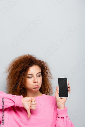 displeased woman pouting lips and showing thumb down near smartphone with blank screen isolated on grey