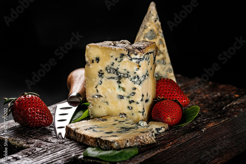 Semi-soft cheese with mold made from cows milk. The king of blue cheeses is Stilton. A piece of cheese and ripe strawberries on a dark wooden board and a cheese knife. Low key image. Copy space