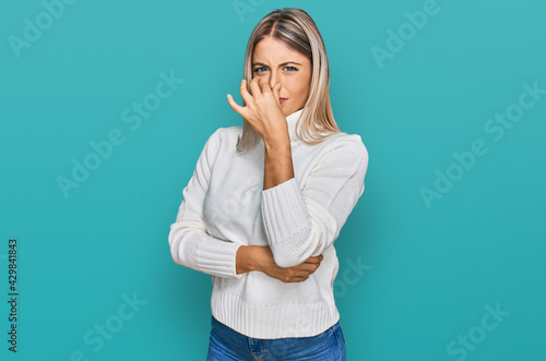 Beautiful blonde woman wearing casual turtleneck sweater smelling something stinky and disgusting, intolerable smell, holding breath with fingers on nose. bad smell