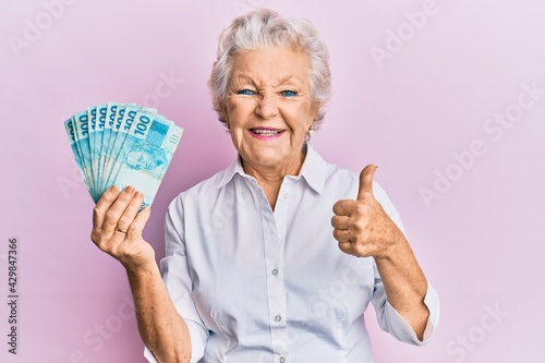 Senior grey-haired woman holding 100 brazilian real banknotes smiling happy and positive, thumb up doing excellent and approval sign