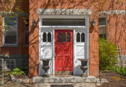 Exterior daytime view of entrance to red brick building showing red entrance door with leaded glass inserts, sidelights and transom window, lamps and planters, nobody