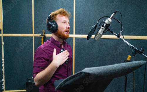 Expressive face of bearded man with red curly hair wear headphones near microphone who makes professional dubbing on a voice recording studio