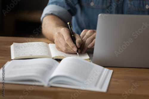 Crop close up of male student make notes handwrite in notebook study online on computer from home. Man write in notepad talking distant on webcam virtual zoom call on laptop. Education concept.