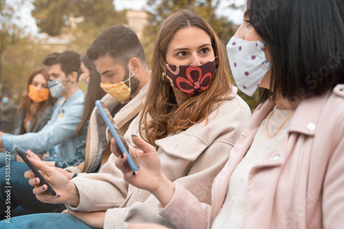 Friends gathering outdoors during coronavirus quarantine talking and hangout together wearing fashion face masks sitting in the park. New normal lifestyle concept.
