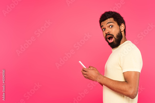 shocked african american man holding smartphone isolated on pink