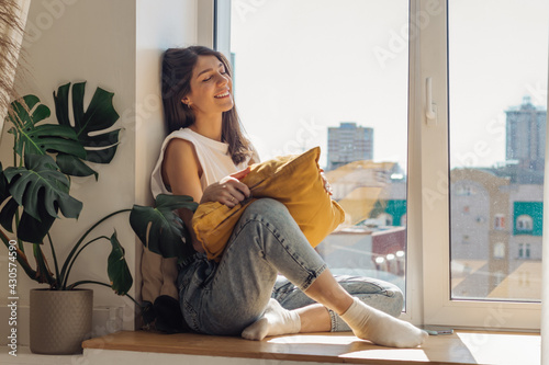 A dark-haired mixed-race girl sitting on the windowsill and enjoying the sunny day.Home interior design.View of the city outside the window.The concept of relaxation and good mood.