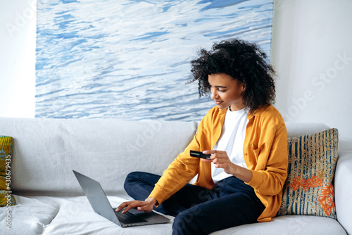 Online shopping concept. Young beautiful african american woman in casual stylish clothes sits on the couch, makes purchases online by laptop, fills in credit card details to pay for goods or delivery