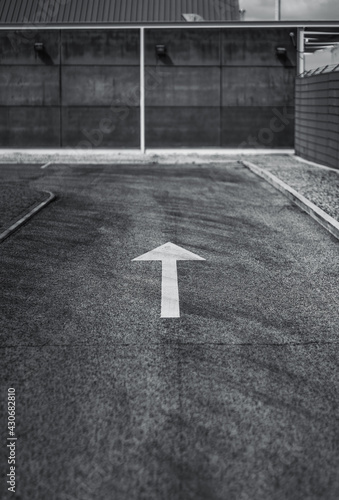 Black and white shot of an asphalt road with a selective focus on a white contrast arrow on it as a road marking leading to a concrete wall and a turn to the left, sunny day, shallow depth of field