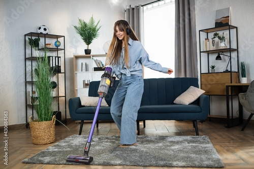 Attractive woman in headphones listening music and doing housekeeping using modern wireless vacuum cleaner. Young housewife enjoying cleaning time with modern technology.