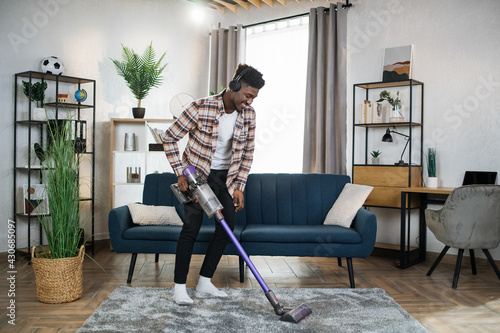 Smiling afro american man in wireless headphones cleaning carpet with handheld vacuum cleaner. Young guy enjoying housekeeping using modern devices.