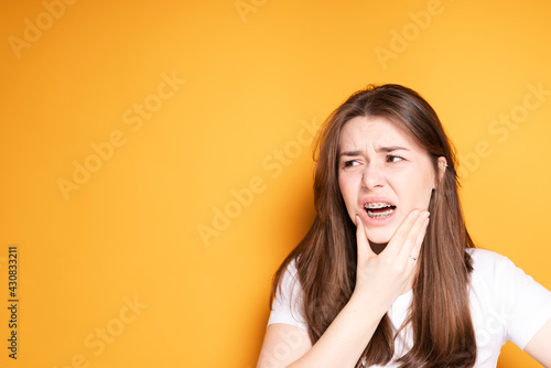 Sad girl in braces touches her jaw suffering from pain in her teeth while standing on a yellow background
