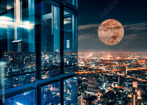 Corner glass window of skyscraper with supermoon glowing on illuminated city