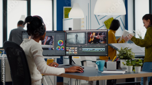 Black videographer putting on headset editing movie using post production software working in creative start up agency office. African editor processing video footage on computer with dual displays