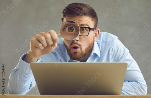 Portrait of funny handsome nerdy young business man in glasses sitting at office desk with laptop computer, holding magnifying glass and looking at something with big eye and surprised face expression