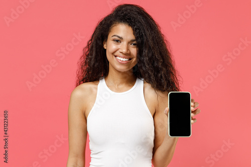 Young fun smiling happy cheerful satisfied african american woman 20s wearing casual white tank shirt hold in hand show mobile cell phone with blank screen workspace area isolated on pink background
