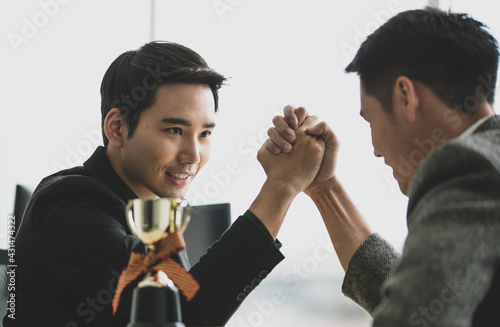 Two young men, one in a black suit, the other in a gray, they playing arm-wrestle for fun, friendly in the office. Noticed from the gentle eyes not aiming to overcome even have a trophies on table.