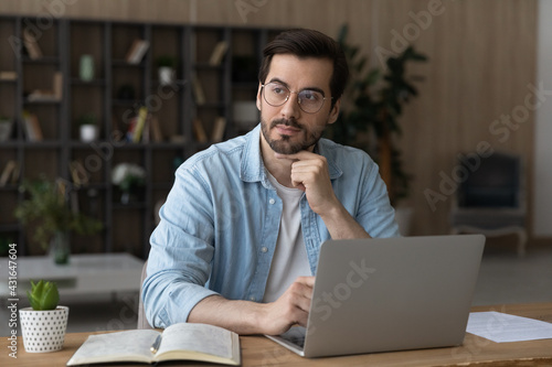 Thoughtful dreamy businessman in glasses sitting at work desk with laptop, looking to aside, pensive young man freelancer or student pondering project strategy, planning, visualizing future