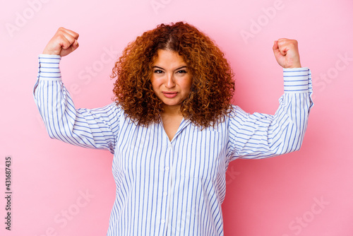 Young latin curvy woman isolated on pink background showing strength gesture with arms, symbol of feminine power