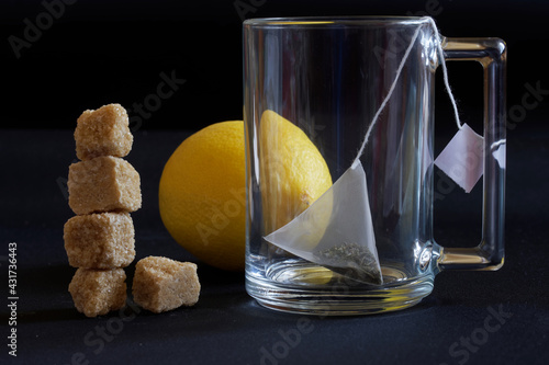 A glass transparent mug with a pyramid tea bag, fresh yellow lemon and a tower of brown cane unrefined sugar cubes. Dark background