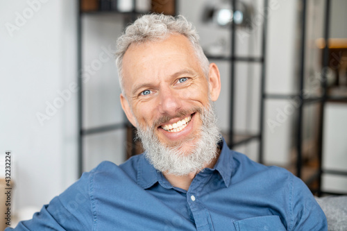 Headshot of handsome charismatic mature man in casual shirt. Close Up portrait of bearded grey-haired middle-aged male, he looks at the camera with a cheerful toothy smile
