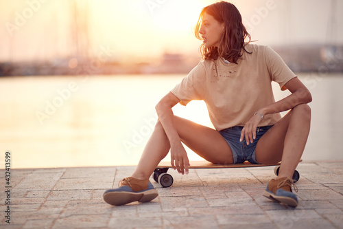 A young beautiful girl is sitting on a skateboard on the dock on the seaside and watching a sunset while taking a rest from a ride. Summer, sea, vacation