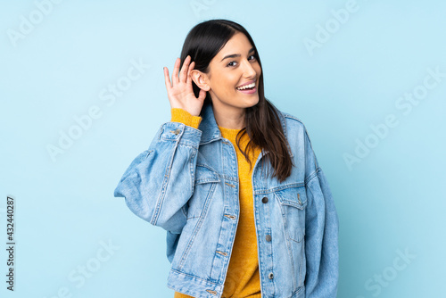 Young caucasian woman isolated on blue background listening to something by putting hand on the ear