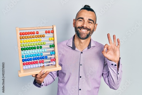 Young hispanic man holding traditional abacus doing ok sign with fingers, smiling friendly gesturing excellent symbol