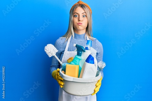 Beautiful blonde woman wearing cleaner apron holding cleaning products puffing cheeks with funny face. mouth inflated with air, catching air.