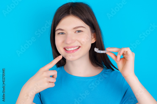 young beautiful Caucasian woman wearing blue T-shirt over blue wall holding an invisible aligner and pointing to her perfect straight teeth. Dental healthcare and confidence concept.