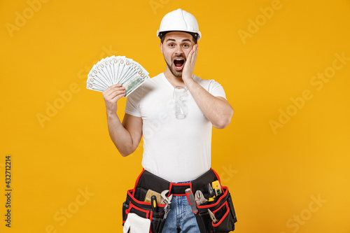Young shocked employee handyman man in protective helmet hardhat hold fan cash money dollar banknotes isolated on yellow background. Instruments accessories renovation apartment room. Repair concept.