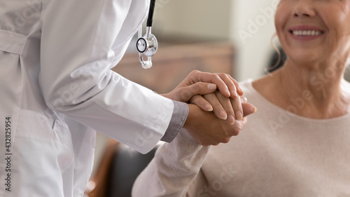 Doctor giving hope. Close up shot of young female physician leaning forward to smiling elderly lady patient holding her hand in palms. Woman caretaker in white coat supporting encouraging old person