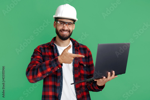 Smiling bearded engineer or constructor man in casual outfit pointing finger on computer over green background.