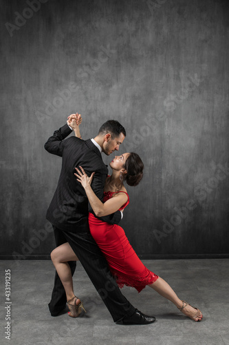Couple of professional dancers in elegant suit and red dress in a tango dancing movement on dark background. Handsome man and woman dance looking eye to eye.