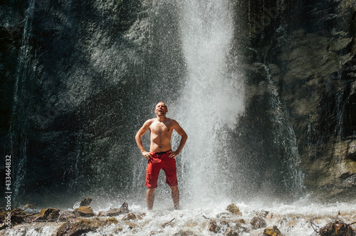 A smiling middle-aged topless man standing under the mountain river waterfall and enjoying the splashing Nature power. Fit people, trekking, and a natural beauty concept image.