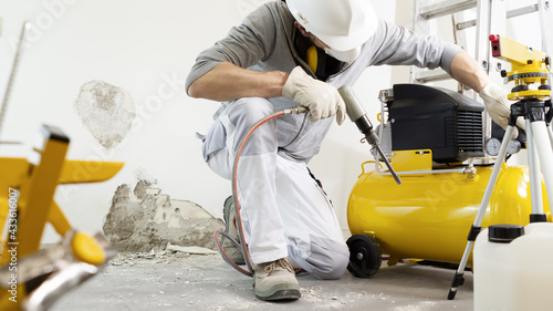 house renovation concept, construction worker breaks the old plaster of the wall with pneumatic air hammer chisel, regulates the airflow of the compressor