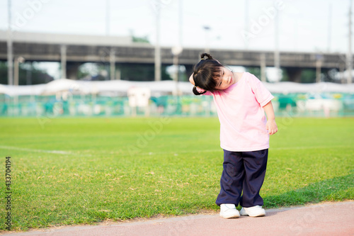 Cute 4 year old Asian girl standing for exercise. Child embraces his arms across the head to opposite side of the head and tilts his neck. Kid enjoy sport. Child wearing pink shirt. Summer or spring.