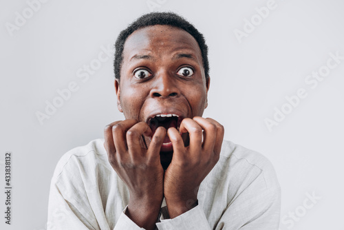 Portrait of a terrified African man staring in horror at the camera and putting his hands to his mouth in a light linen shirt on a white background. Very strong surprise or fright, horror in the eyes.