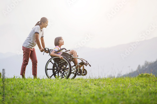 Disabled handicapped child sitting in wheelchair and care helper walking on mountain meadow park in sunny day.. International Disability Day.