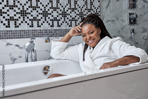 Adorable woman with perfect dark skin sits inside of bathtub, poses in bathroom enjoying weekends, self-care, romantic time alone