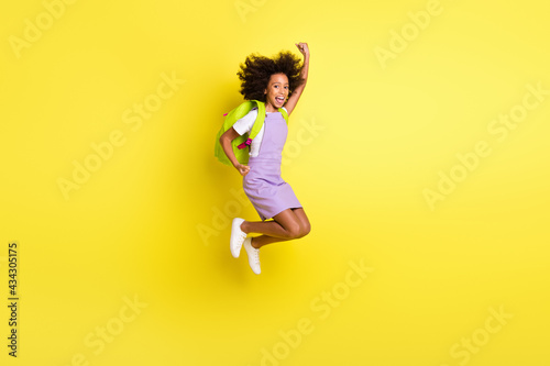 Full length body size photo of funny schoolgirl jumping high gesturing like winner wearing backpack isolated bright yellow color background