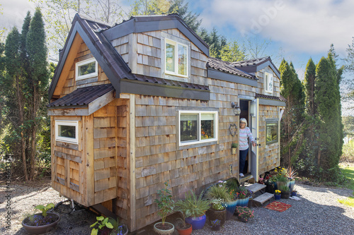 Friendly mature woman standing at front door of her charming little wooden tiny home or house