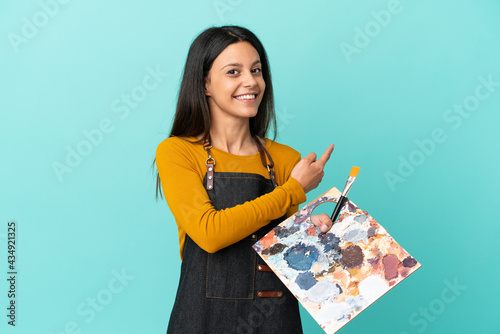 Young artist caucasian woman holding a palette isolated on blue background pointing back