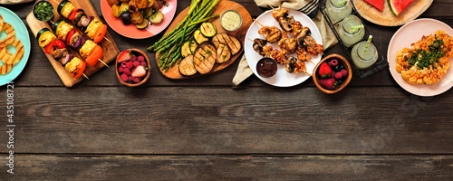 Healthy plant based summer bbq top border. Above view over a dark wood banner background. Grilled fruit and vegetables, skewers, cauliflower steak and vegetarian sides. Copy space.