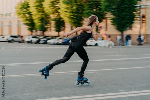 Professional female roller demonstrates her abilities of rollerblading rides very quckly on road along city enjoys sunny day dressed in black active wear. Active lifestyle hobby and fitness activity