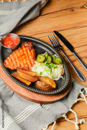 Juicy grilled pork steak with baked potatoes, cut pickles and tomato sauce served in a tray over rustic wooden table.