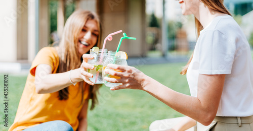  Two beautiful young women sitting on the grass and laughing while having a lemonade cocktails drink in glass jars during a friendly hang out in city park. Summer vacation and picnic at sunny day.