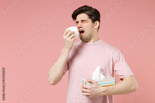 Sick unhealthy ill allergic man has red watery eyes runny stuffy sore nose suffer from allergy trigger symptom hay fever hold paper napkin handkerchief isolated on pastel pink color background studio