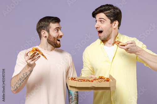 Two excited young men friends together in casual t-shirt tattoo translate fun eat italian pizza in cardboard flatbox look to each other isolated on purple background studio People lifestyle concept.