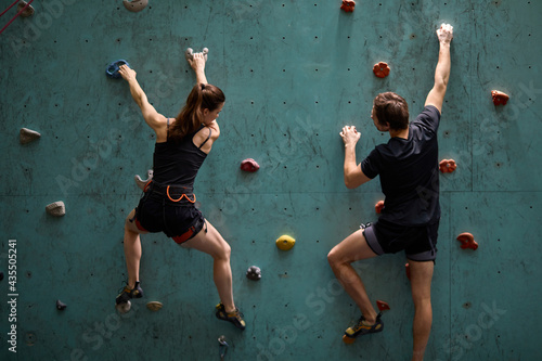 Professional woman and man mountaineers climbing artificial rock wall with belay at bouldering gym, in sportswear, rear view on professional sport people workout training